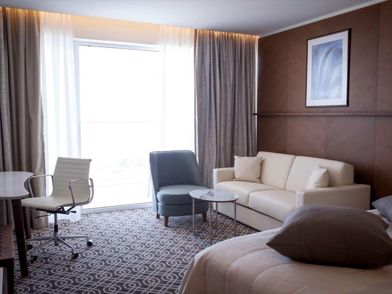 Moderně zařízený hotelový pokoj s postelí, pohovkou, židlemi a velkým oknem.
