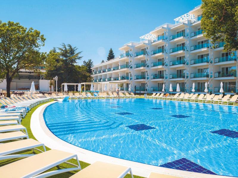 Velký hotel s čistým bazénem a lehátky pod stromy za jasné oblohy.