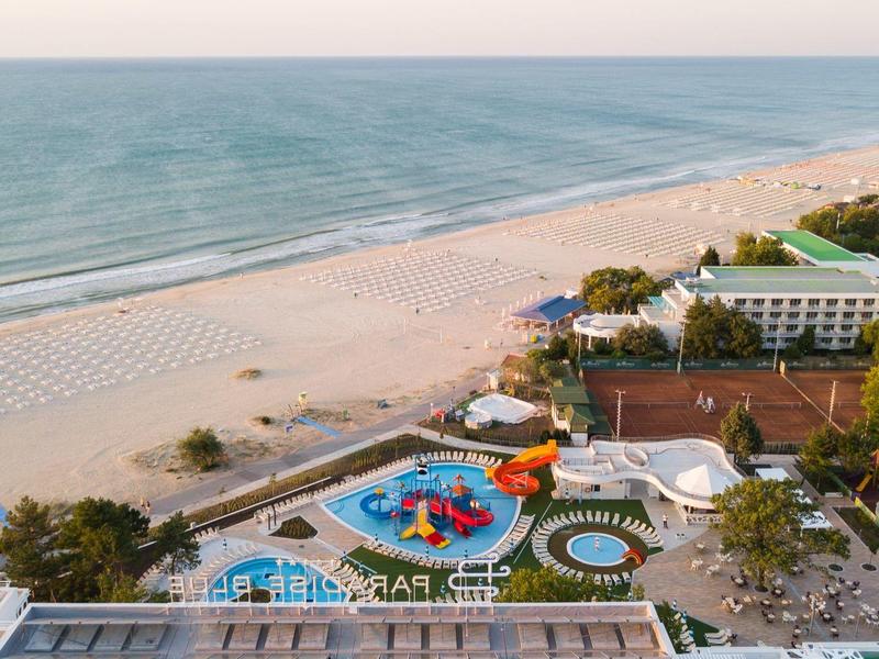 Pohled na pláž s přilehlým hotelem a barevným vodním parkem za jasné oblohy.