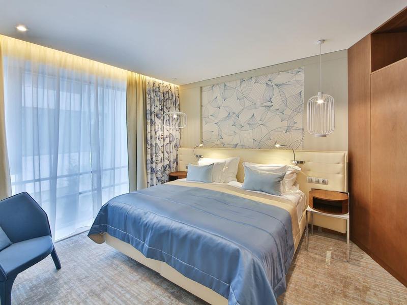 Moderně zařízený hotelový pokoj s velkou postelí, modrou přikrývkou a balkonovým oknem.