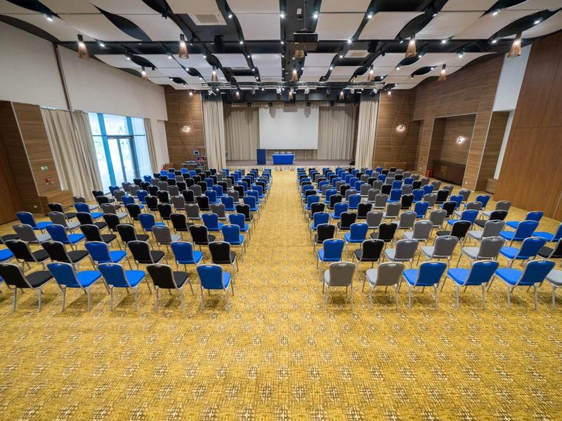 Velký konferenční sál s řadami modrých a černých židlí na žlutém koberci.