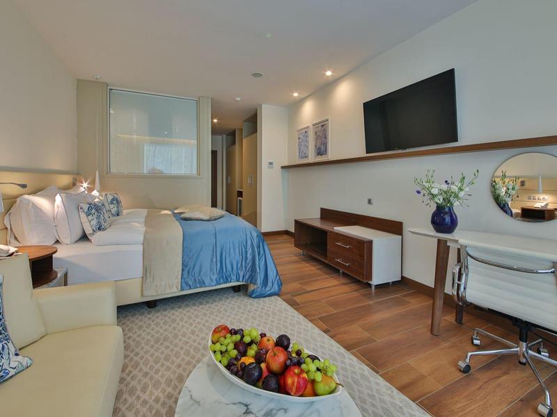 Moderní hotelový pokoj s manželskou postelí, pracovním stolem, televizí a kancelářskou židlí.