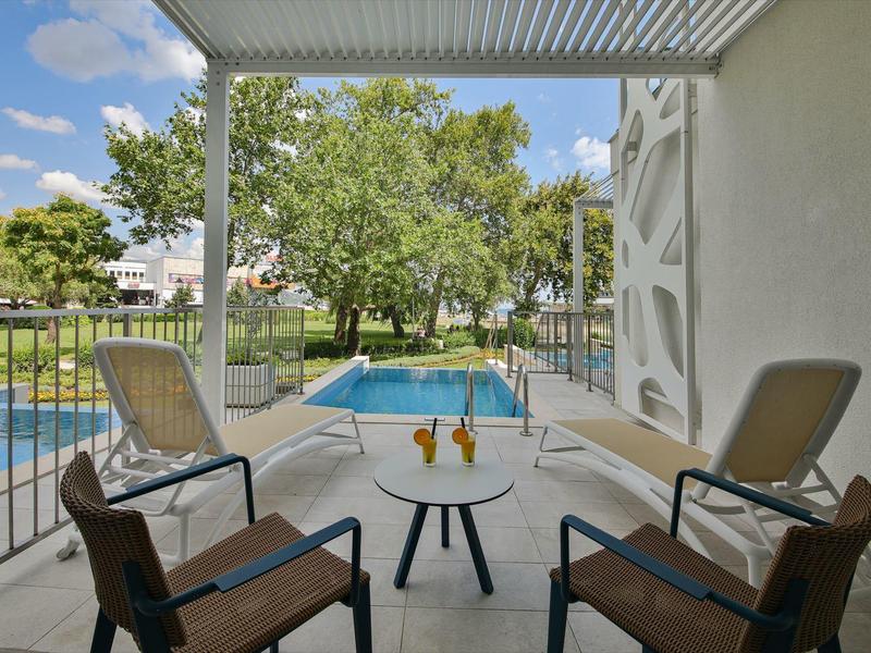 Balkon s posezením s výhledem na bazén a stromy pod modrou oblohou.