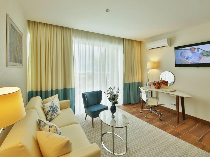 Světý hotelový pokoj s posezením, závěsy a televizí na stěně.