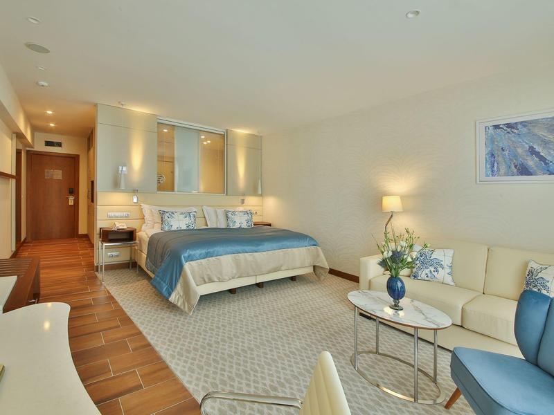 Světý hotelový pokoj s manželskou postelí, pohovkou, křesly a moderním zařízením.