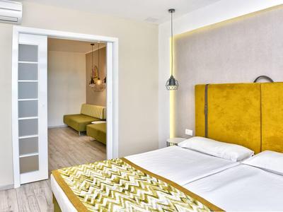 Habitación de hotel moderna con dos camas, cabecero amarillo y acceso a una sala contigua con área de estar.