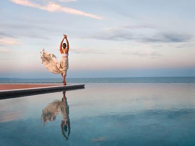 Mujer bailando elegantemente al borde de la piscina con mar y cielo tranquilos al fondo.
