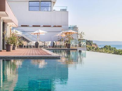 Modern zwembad met terras en witte parasols, naast zeezicht bij heldere lucht.