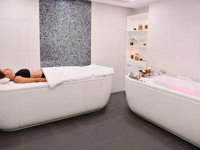 Mujer relajándose en una camilla blanca junto a una bañera con agua rosa en un spa moderno.