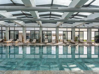 Piscina interior moderna con tumbonas en un área luminosa de hotel con techo de vidrio y fachada de ventanas.