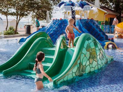 Niños juegan en tobogán verde en área poco profunda de piscina con diseño de rocas.