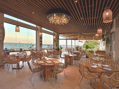 Terraza abierta de hotel con techo de madera, mesas redondas y vista al mar al atardecer.