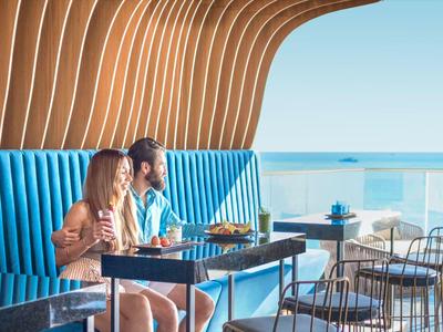 Zwei Personen sitzen in einem modernen Restaurant mit Meerblick und blauen Sitzbänken.