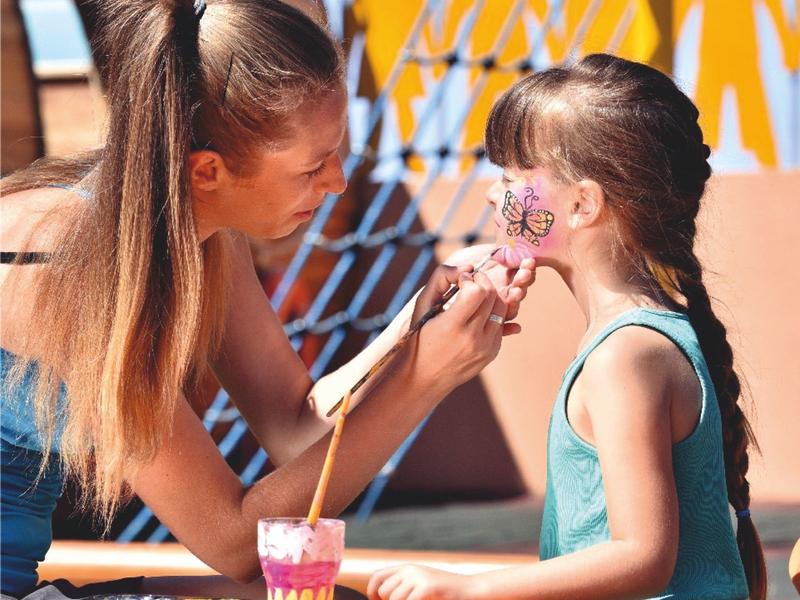 Una niña recibe pintura facial de una mujer en un entorno soleado de viaje o actividad vacacional.