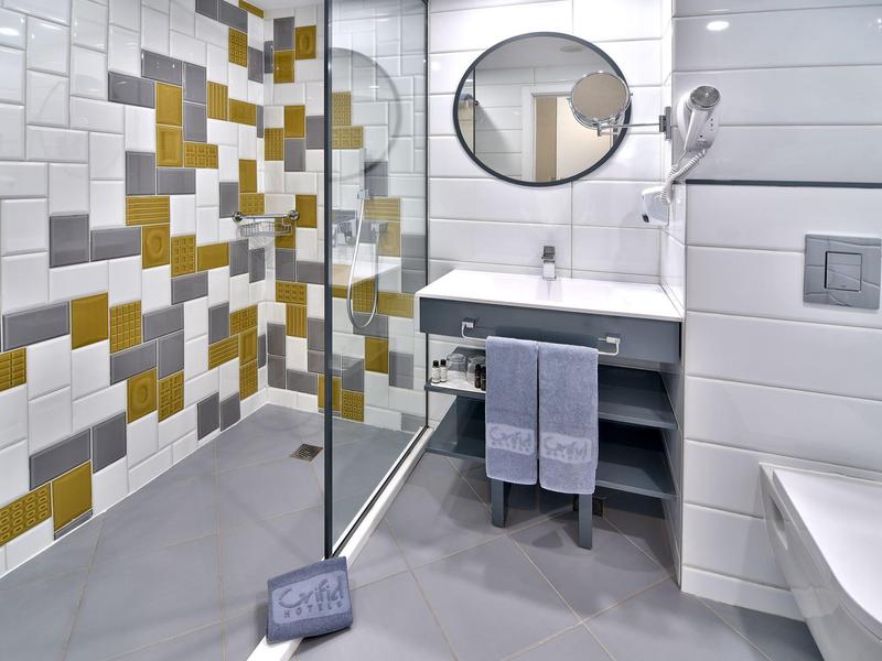 Baño moderno con pared de azulejos coloridos, mampara de vidrio, lavabo y espejo redondo.