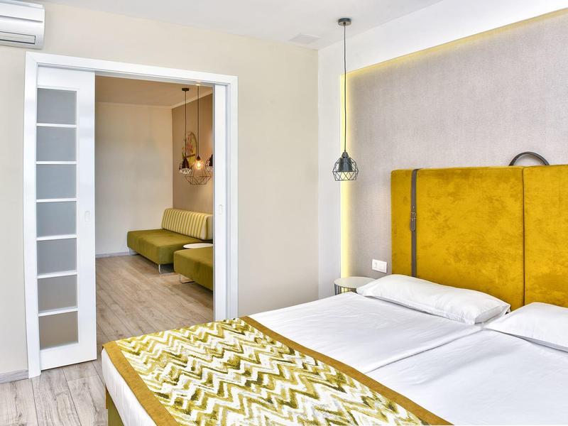 Habitación de hotel moderna con dos camas, cabecero amarillo y acceso a una sala contigua con área de estar.