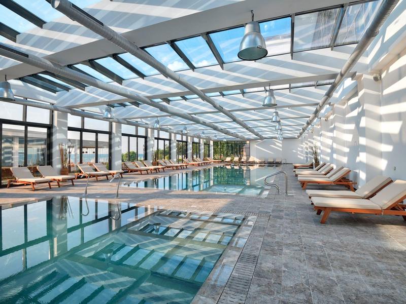 Área moderna de piscina interior con tumbonas y techo de cristal en ambiente luminoso.