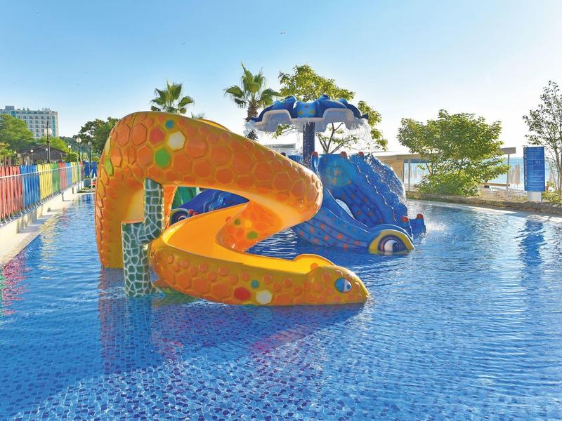 Área de juegos acuáticos colorida con toboganes de jirafa y delfín en la piscina bajo cielo azul.