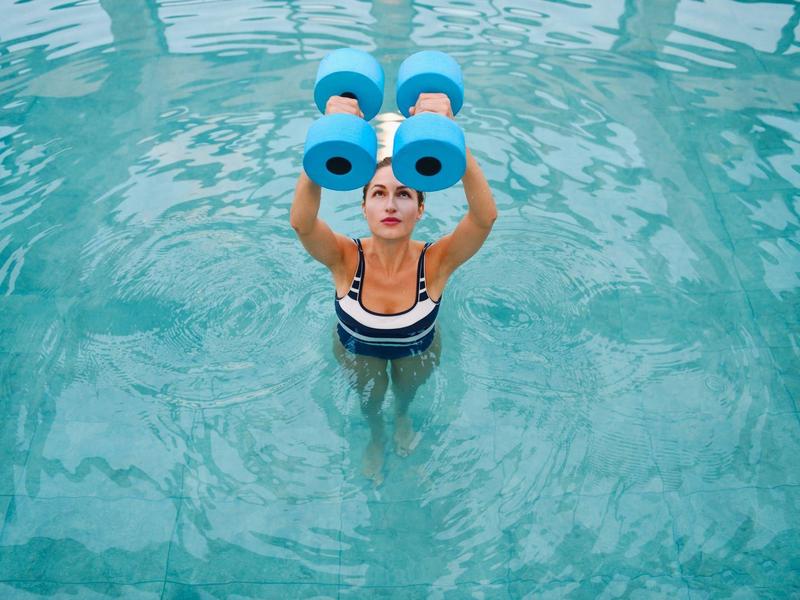 Mujer en la piscina sostiene pesas de espuma azules sobre el agua durante entrenamiento acuático.