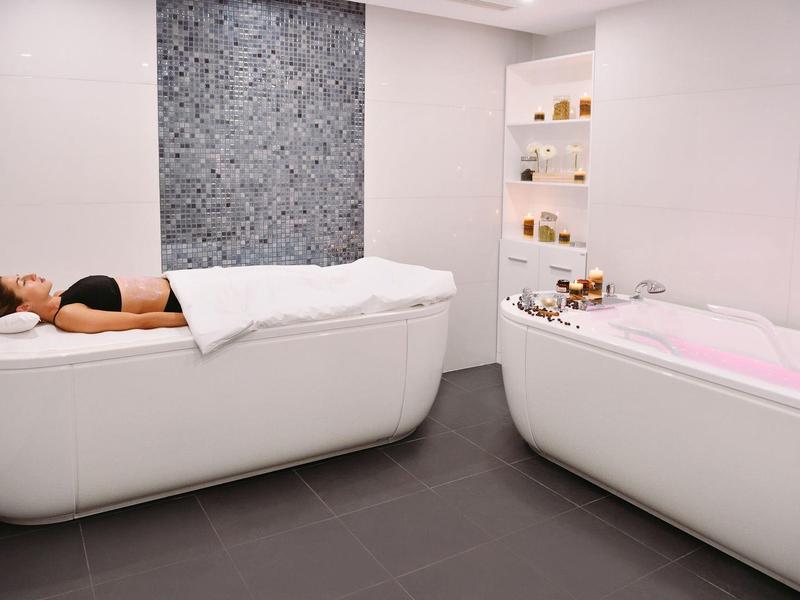 Mujer relajándose en una camilla blanca junto a una bañera con agua rosa en un spa moderno.