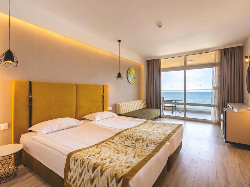 Habitación de hotel iluminada con cama doble y vista al mar a través de grandes ventanales.