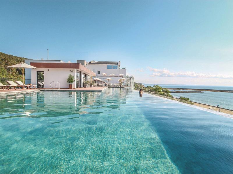 Piscina infinita con vista al mar junto a modernos edificios de hotel blancos bajo cielo azul.