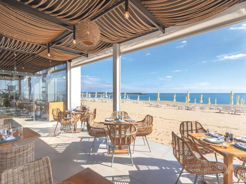 Restaurante abierto en la playa con vista a la arena, sombrillas azules y mar tranquilo.