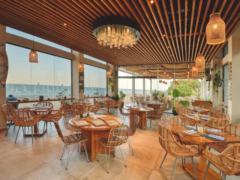 Terraza luminosa de hotel con mesas de madera y sillas de alambre, vista al mar durante el día.