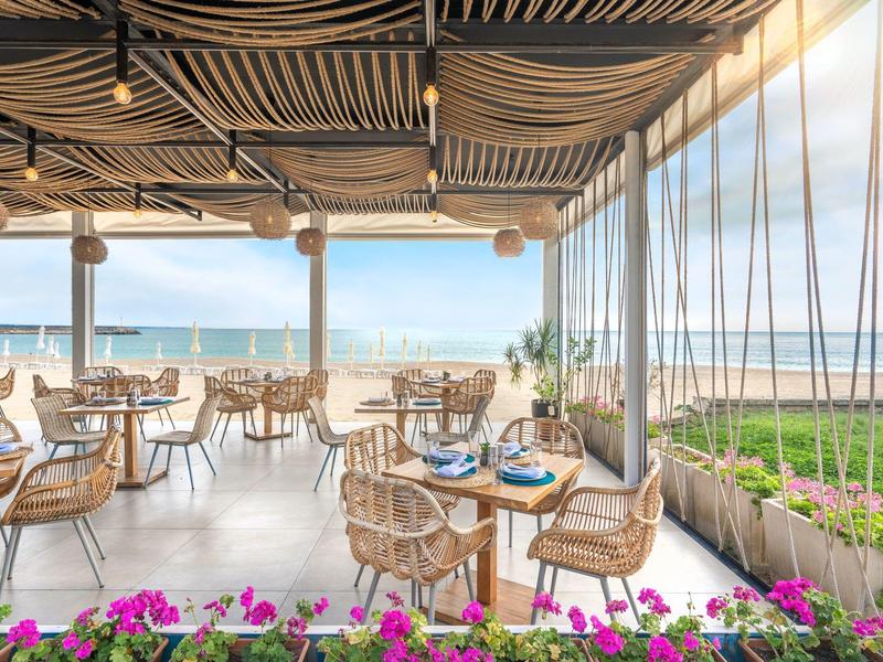 Área de restaurante al aire libre con sillas de mimbre y vista al mar y la playa en cielo despejado.