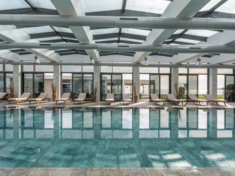 Piscina interior moderna con tumbonas en un área luminosa de hotel con techo de vidrio y fachada de ventanas.