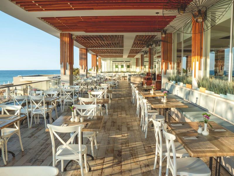 Restaurante abierto con sillas blancas y vista al mar al atardecer.