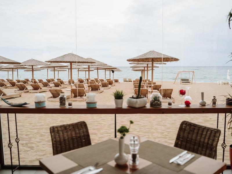 Vista de una terraza en la playa con mesas puestas, sillas y sombrillas en la arena.