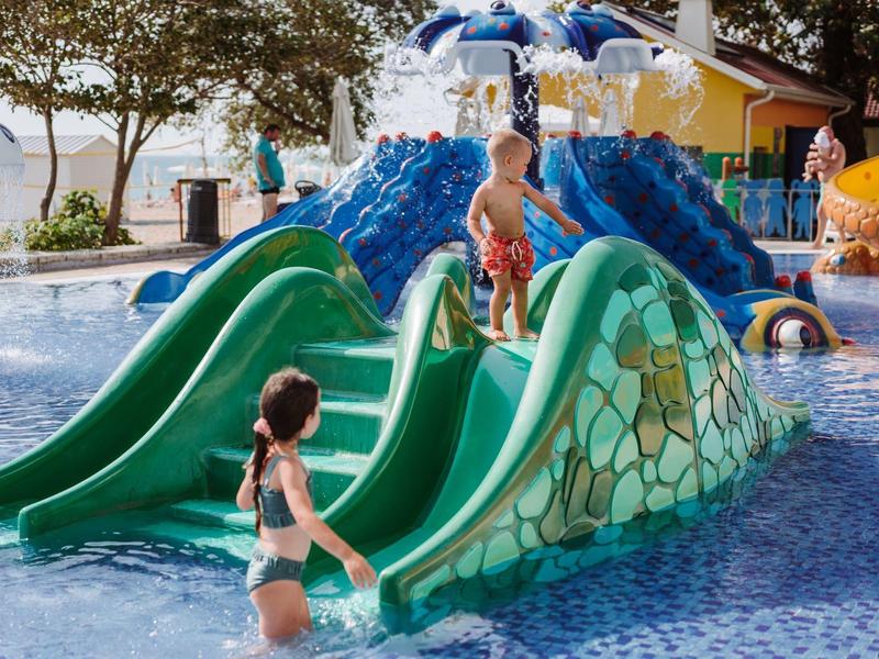 Niños juegan en tobogán verde en área poco profunda de piscina con diseño de rocas.
