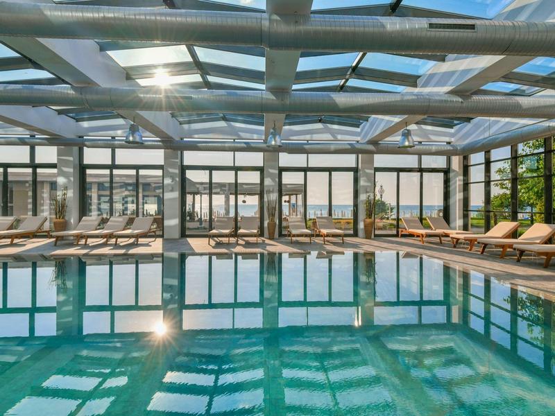 Piscina cubierta con tumbonas y techo de cristal, mucha luz solar y ambiente moderno.