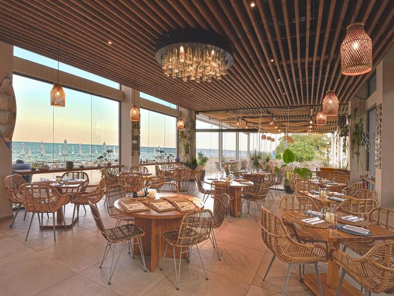 Terraza abierta de hotel con techo de madera, mesas redondas y vista al mar al atardecer.