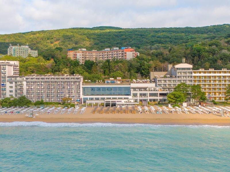 Hotel de playa con varios edificios frente a colina boscosa y mar tranquilo en primer plano
