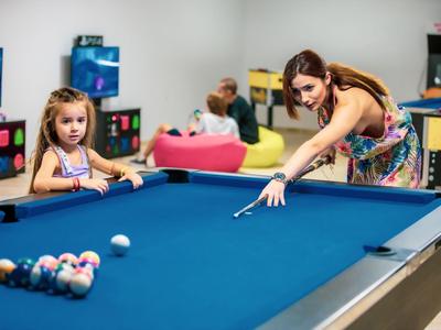 Frau spielt Billard an blauem Tisch, Kind beobachtet, belebter Raum mit Sitzbereich im Hintergrund.