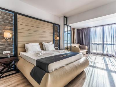 Habitación de hotel moderna con cama doble grande, zona de estar y ventanas del suelo al techo con cortinas.