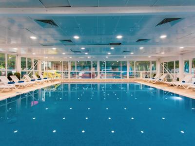 Piscina cubierta con agua tranquila, rodeada de tumbonas blancas y grandes ventanas.