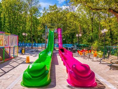 Colorida zona de juegos infantiles con toboganes verde y rosa en un área soleada con bancos.