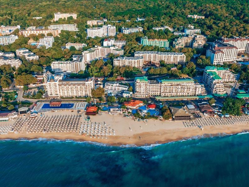 Vista aérea de un gran hotel de playa con muchas tumbonas y entorno verde a lo largo de la costa.