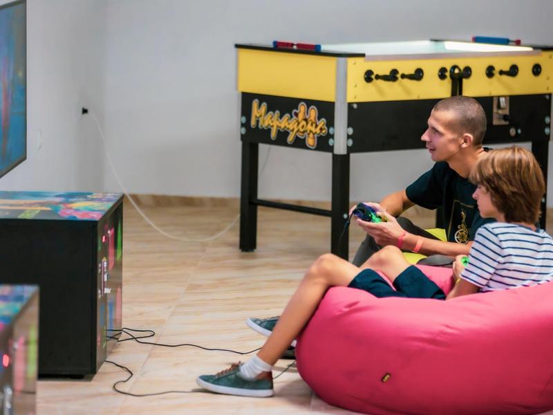 Dos niños sentados en un puff juegan videojuegos junto a una mesa de futbolín.