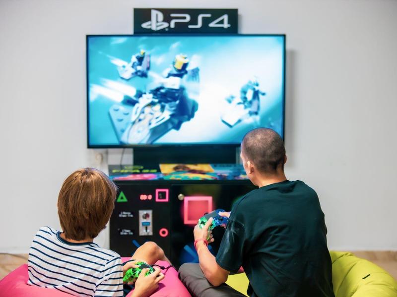 Dos personas juegan videojuegos en una PlayStation 4 frente a una gran televisión en la sala.