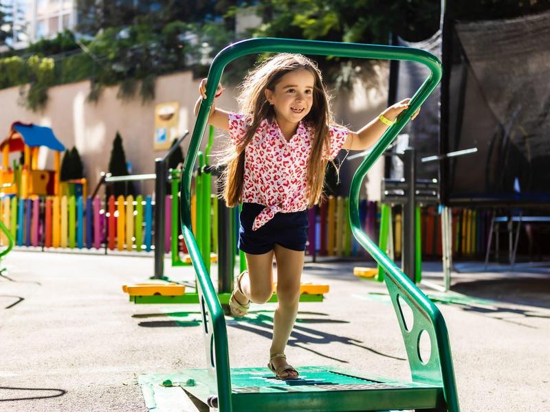 Una niña juega al aire libre en una estructura de escalada verde en un parque infantil con sol.