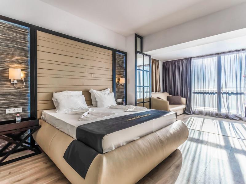 Habitación de hotel moderna con cama doble grande, zona de estar y ventanas del suelo al techo con cortinas.