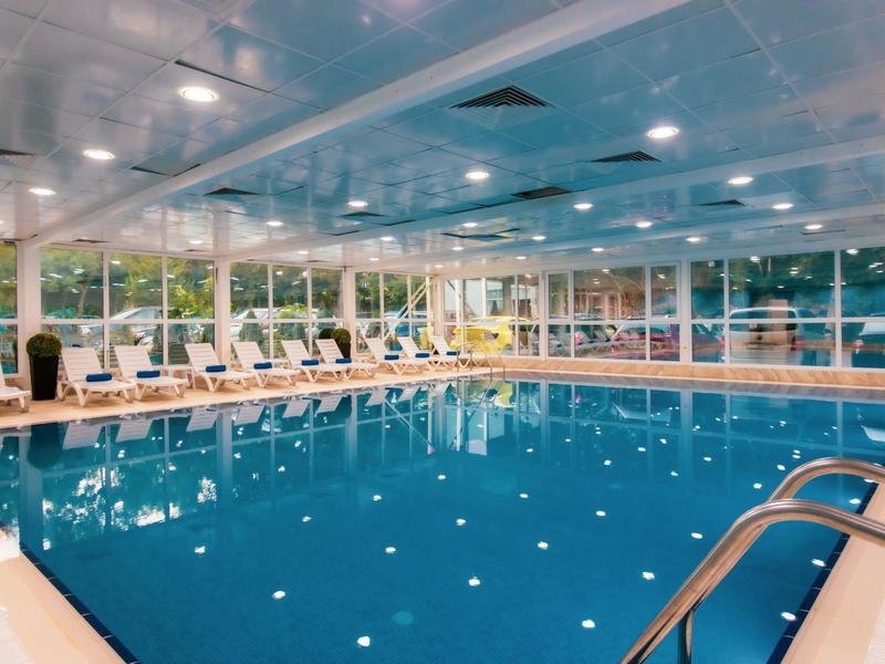 Gran piscina cubierta con tumbonas y ventanas panorámicas con vista a un jardín.