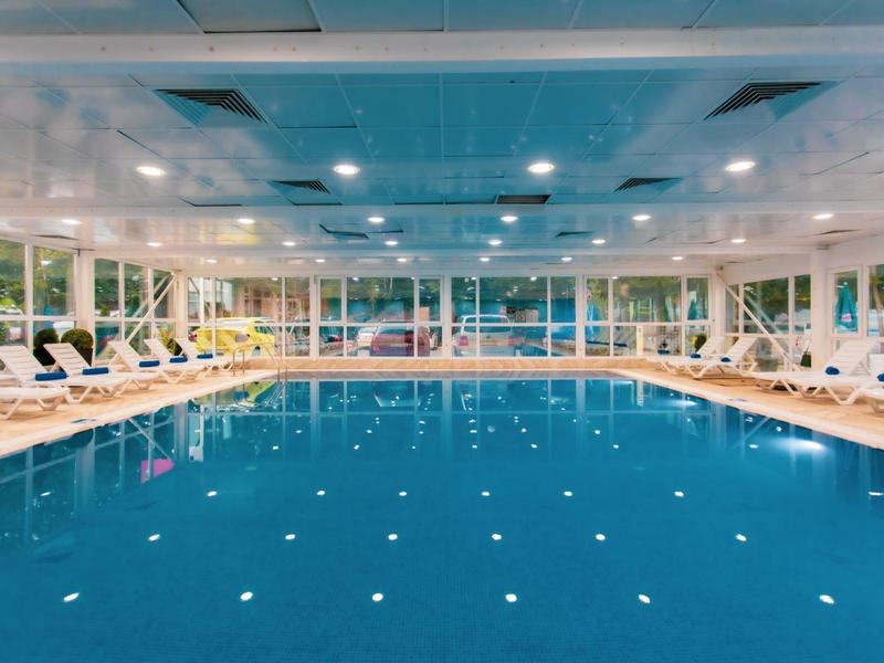 Piscina cubierta con agua tranquila, rodeada de tumbonas blancas y grandes ventanas.