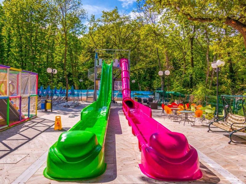 Colorida zona de juegos infantiles con toboganes verde y rosa en un área soleada con bancos.