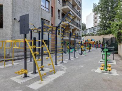 Buitenfitnessruimte met trainingsapparatuur naast een woongebouw onder bomen.