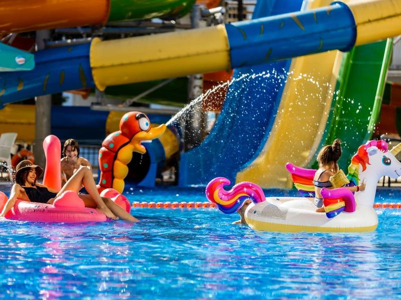 Twee vrouwen ontspannen op roze en witte eenhoorn zwembandjes in het zwembad naast kleurrijke glijbanen.